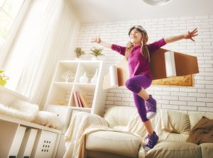 A young girl wearing a purple outfit and aviator goggles joyfully plays with cardboard wings attached to her back, standing on a light-colored couch in a bright, cozy living room. She has her arms spread wide, pretending to fly.