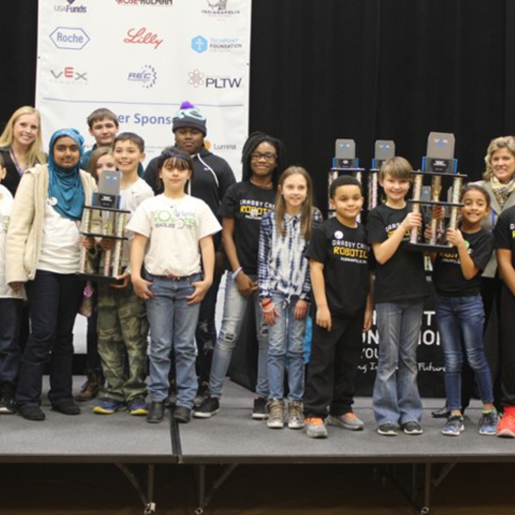 A group of young students stands on a stage, some holding trophies, after a robotics competition. They are accompanied by a few adults for a group photo. Behind them is a banner displaying sponsor logos.