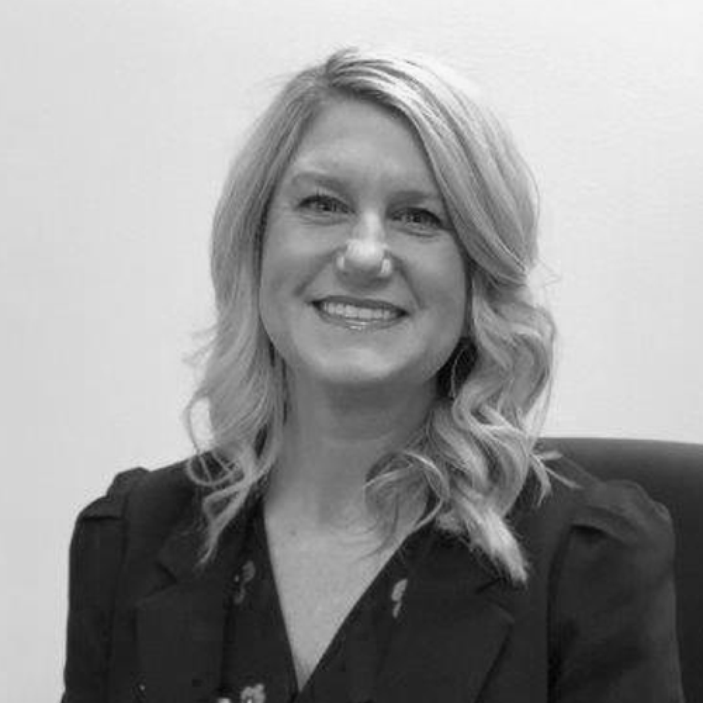 A person with shoulder-length, wavy hair is smiling, wearing a dark blazer and seated in front of a plain, light-colored background. The photo is in black and white.