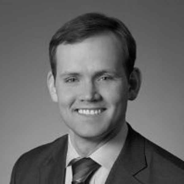 Black and white portrait of a man wearing a suit and tie, smiling at the camera. The background is plain and neutral, directing focus to his face. The overall demeanor appears professional and approachable.