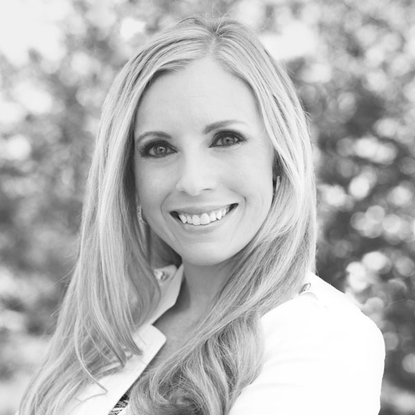 A woman with long hair smiles while looking at the camera. She is wearing a white jacket, and the background is blurred with trees. The image is in black and white.