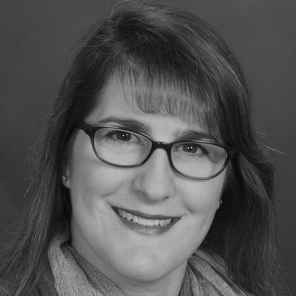 A black-and-white portrait of a person with shoulder-length hair and glasses, smiling at the camera. They are wearing a scarf and have straight bangs just above the eyebrows. The background is plain and dark, keeping the focus on their face.