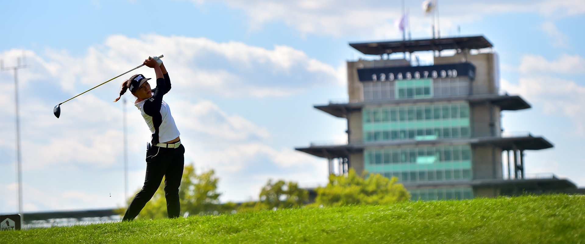 A golfer finishes a powerful swing on a lush green course with a large, multi-tiered building in the background under a partly cloudy sky.