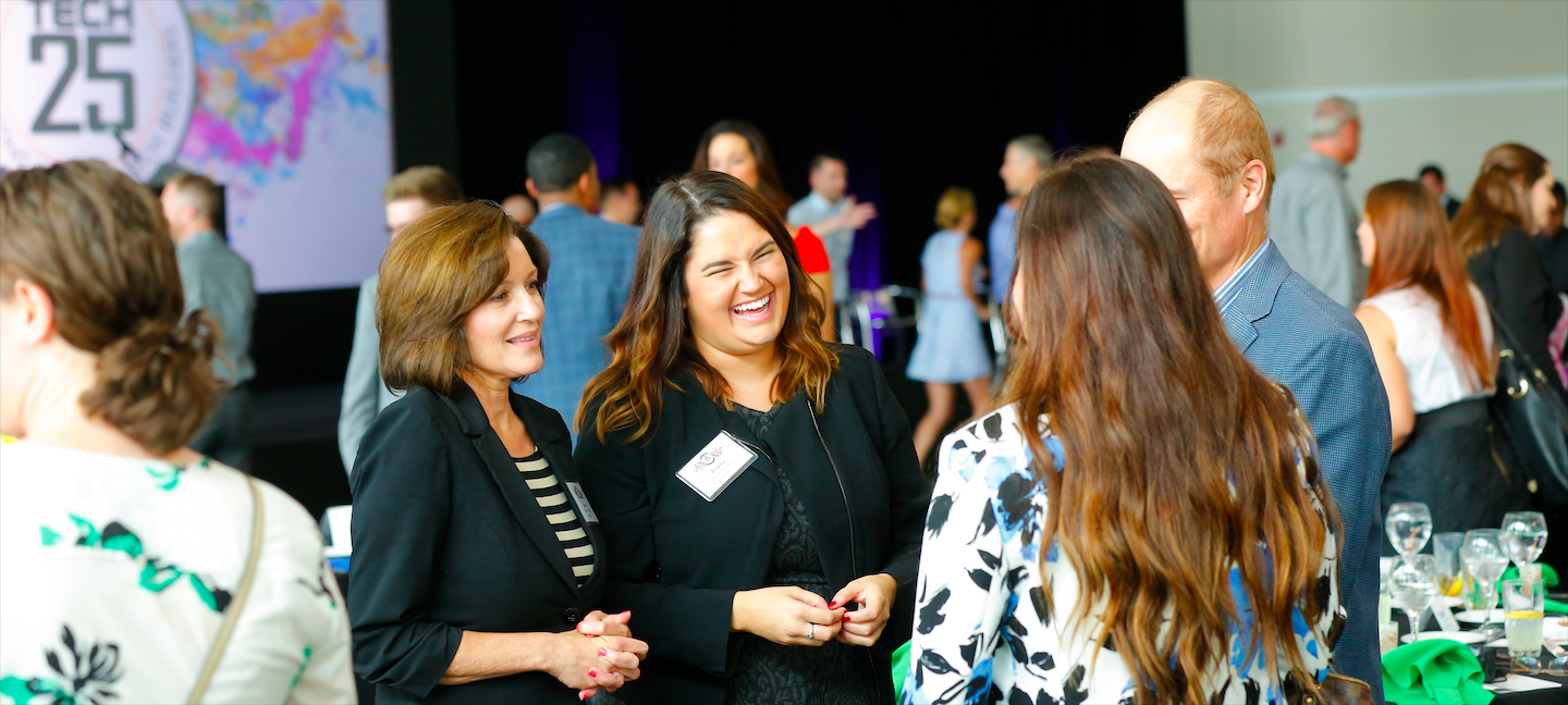 A group of people interact at an indoor networking event with a "Tech 25" sign in the background. They are dressed in business attire and appear to be engaged in lively conversation. Tables with wine glasses and plates are visible.