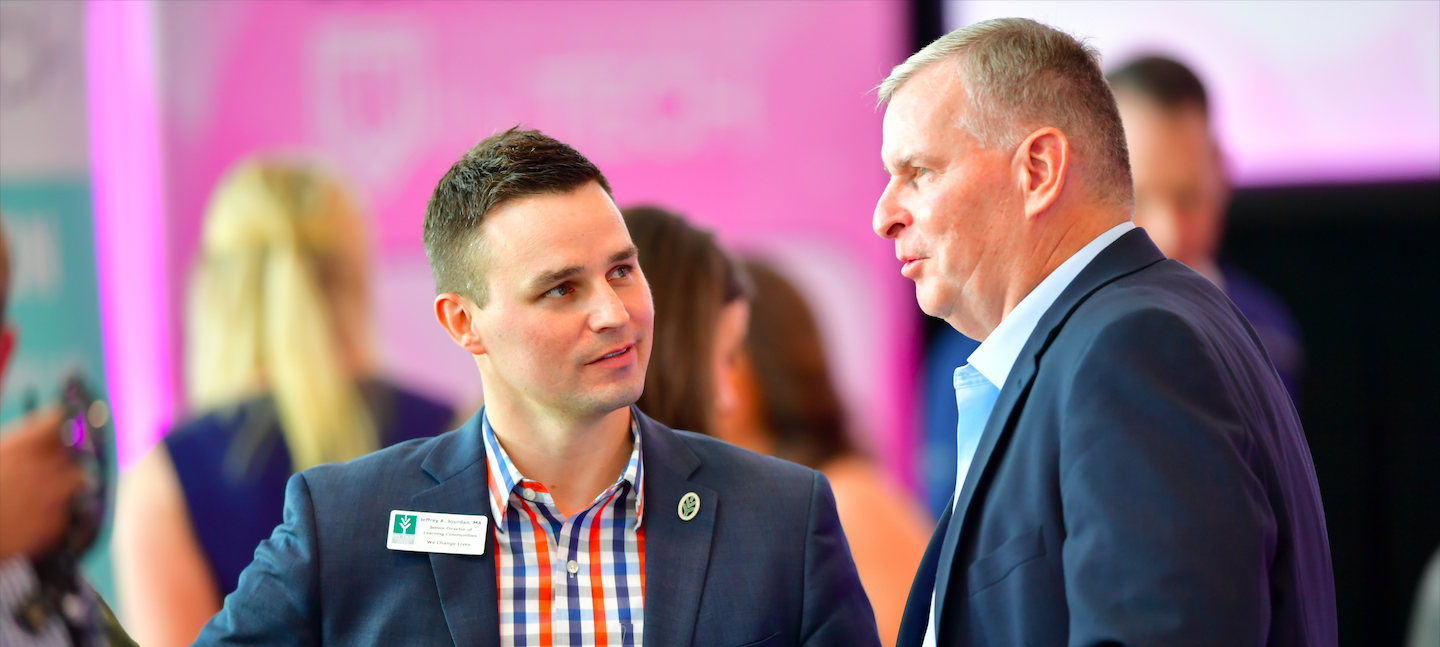 Two men, both in suits, engage in conversation at a networking event. One, wearing a plaid shirt under his blazer, has a name badge. The background is blurred with people and a pink-lit ambiance.