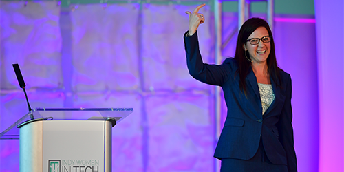 A woman in a navy blue suit with glasses is speaking on stage, gesturing with one hand. She stands next to a podium with a microphone and a sign that reads "INDY WOMEN IN TECH." The stage backdrop is lit with purple and blue lights.