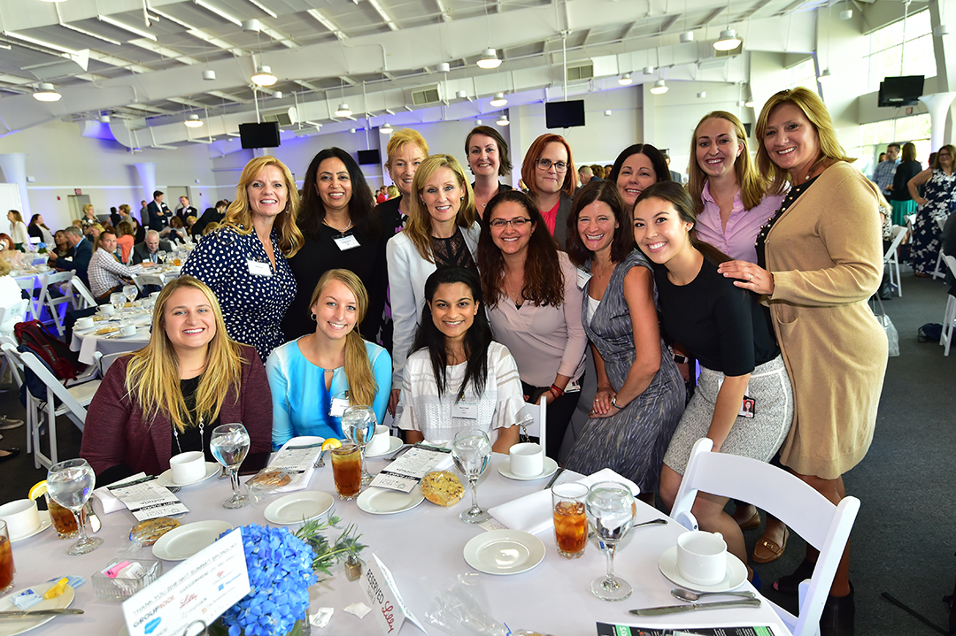 A group of smiling women gather around a table at a formal event in a bright, spacious venue. Some are standing while others are seated. The table is set with plates, drinks, and programs. The background shows other attendees and tables, suggesting a large gathering.