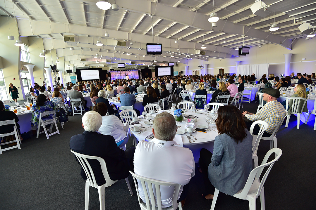 A large group of people sits at round tables during a formal event in a spacious, well-lit hall. Attendees face a stage with presentation screens. The setting includes white chairs and tables, with some guests engaged in conversation.