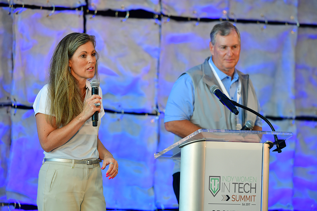 A woman and a man stand on stage at a podium labeled "Indy Women in Tech Summit." The woman speaks into a microphone while holding her other hand at her side, and the man stands behind the podium, listening attentively. The background features a blue, quilt-like design.