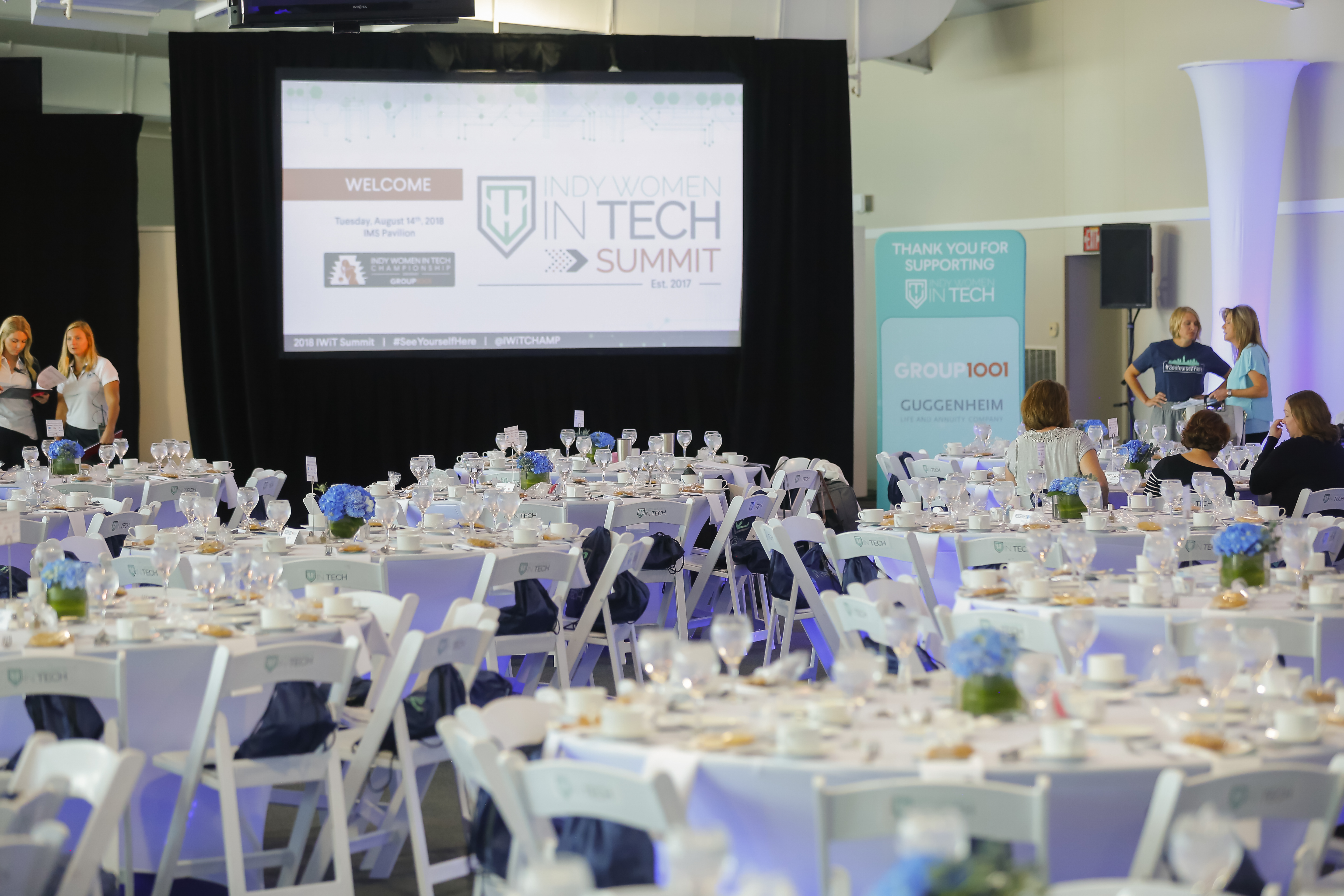 A spacious event hall is set up with numerous round tables adorned with white tablecloths and simple floral centerpieces. A large screen displays "Indy Women in Tech Summit" info. People are visible in the background near a podium and sponsor banners.