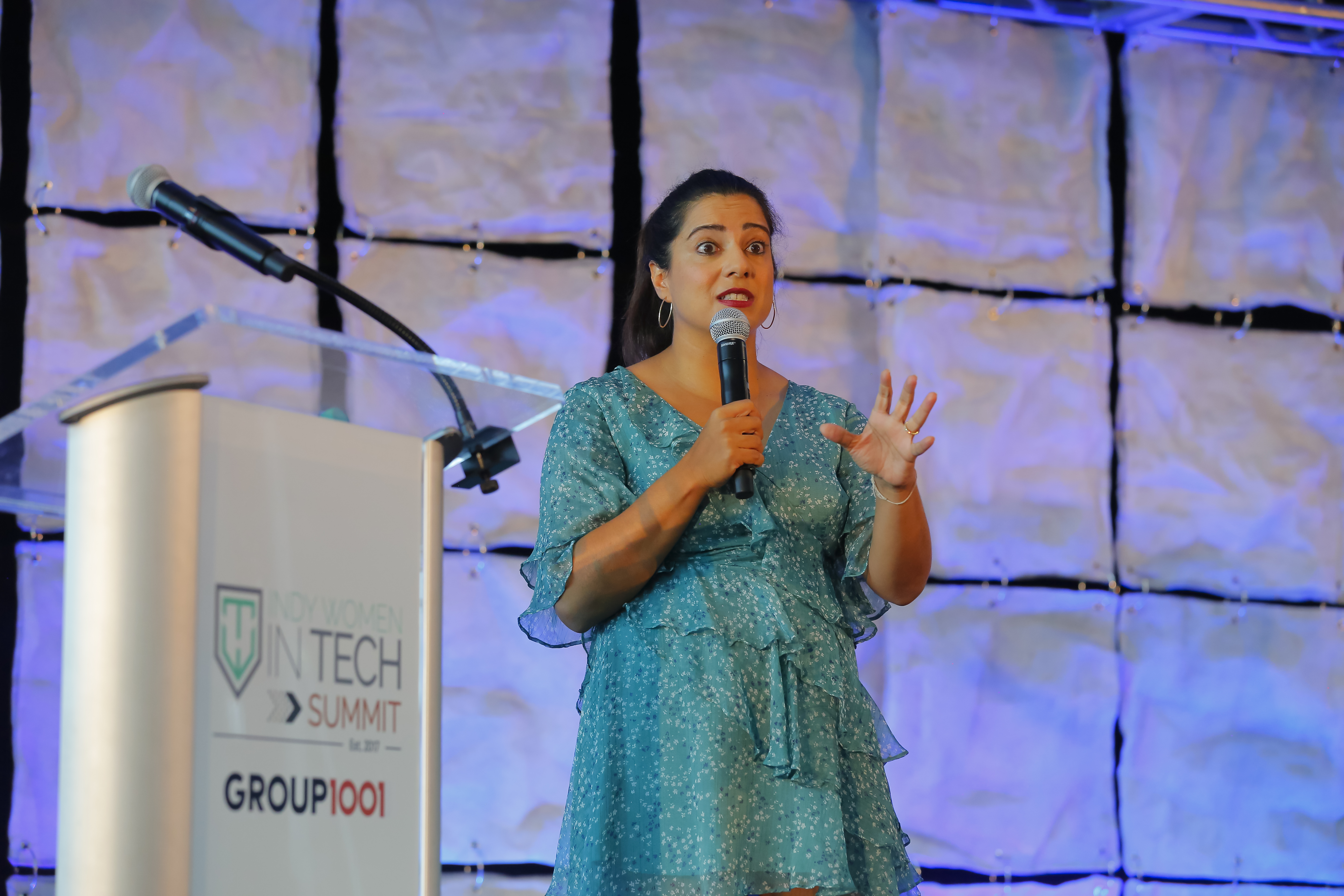 A woman in a blue dress is speaking into a microphone on stage at the Women in Tech Summit. She is standing next to a clear podium with the event's logo and a backdrop featuring a textured design.