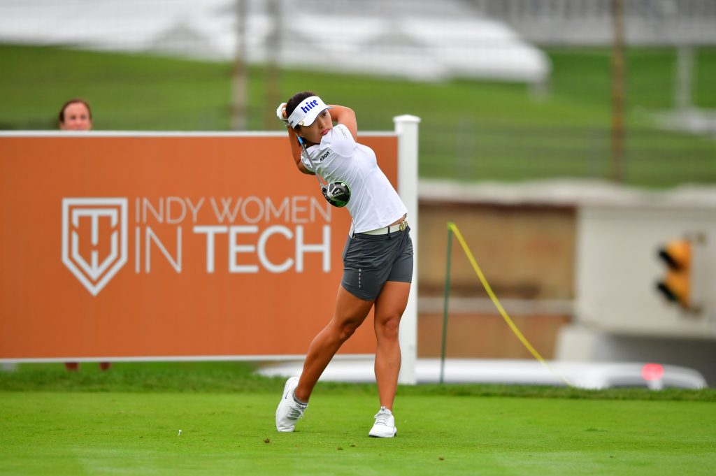 A golfer wearing a white shirt and cap, gray shorts, and white shoes swings a club on a green field at the "Indy Women in Tech" event. In the background, another person watches near a large orange sign with the event's logo and name.