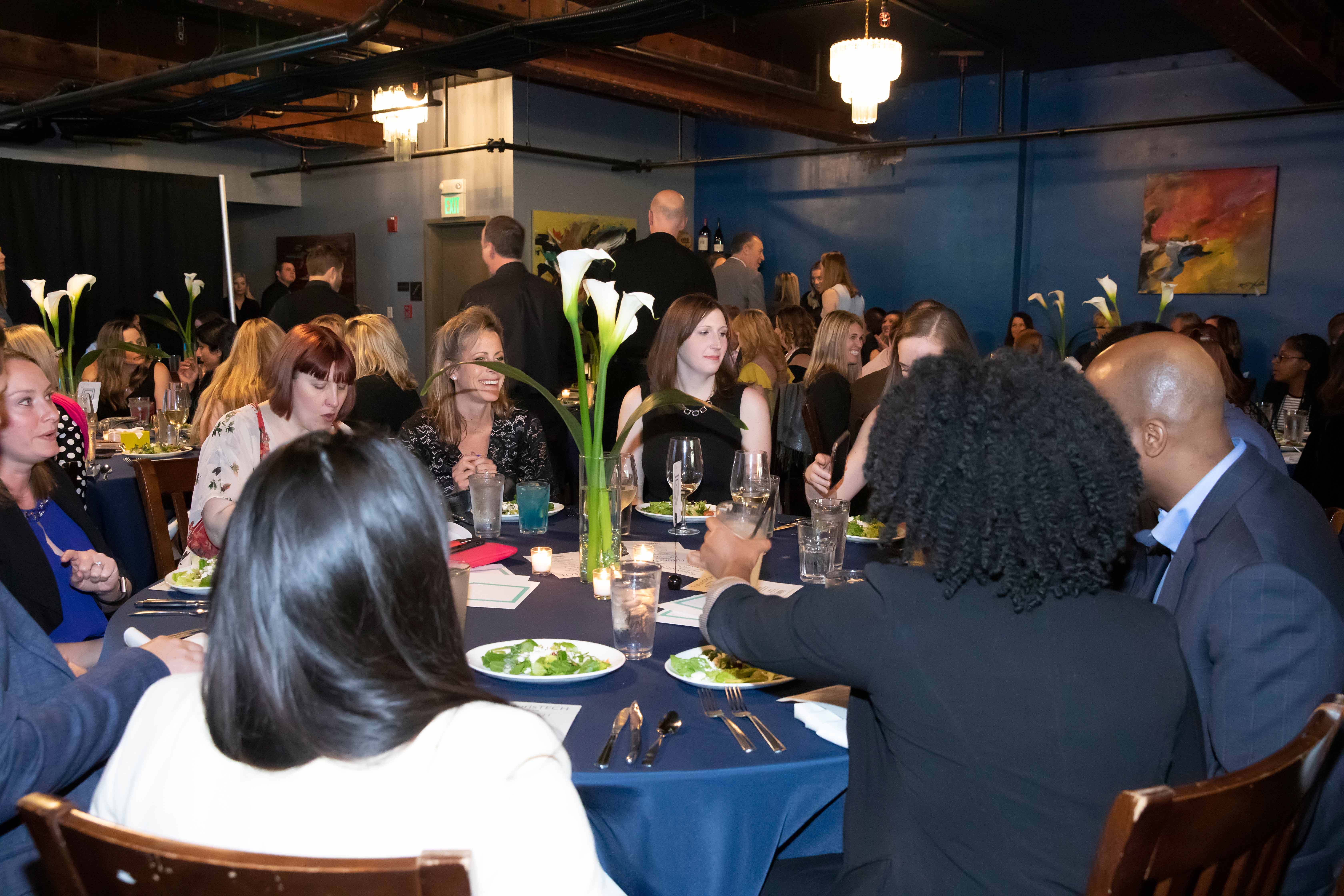 A group of people dressed formally are seated around a round table at an event, engaging in conversation. The table is set with plates of salad, utensils, and tall vases with white flowers. The background features more tables, attendees, a blue wall, and paintings.