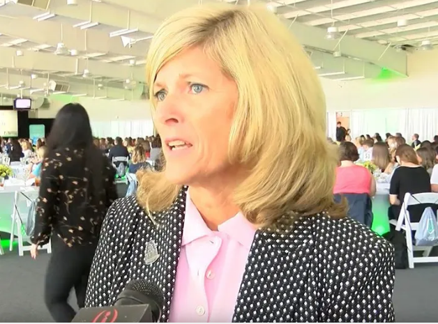 A woman with blonde hair, dressed in a white blouse and a dark polka-dotted blazer, is speaking in an indoor event venue. She is seen from the shoulders up, with a microphone in front of her. The background is blurry, showing several people seated at tables.